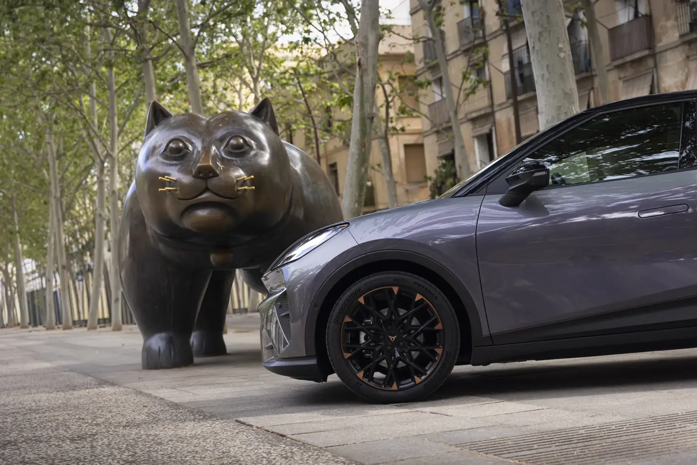 En grå CUPRA Raval holder parkeret ved siden af en stor bronzeskulptur af en kat i en allé med træer og bygninger i baggrunden.