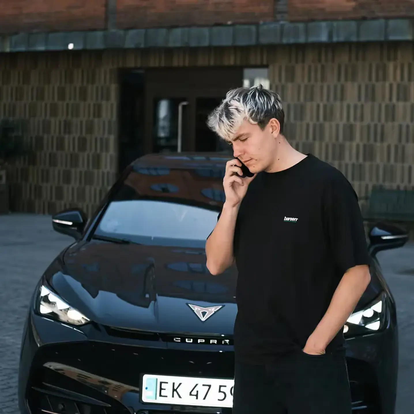 Ung mand taler i telefon foran en sort CUPRA-bil parkeret i et bymiljø.