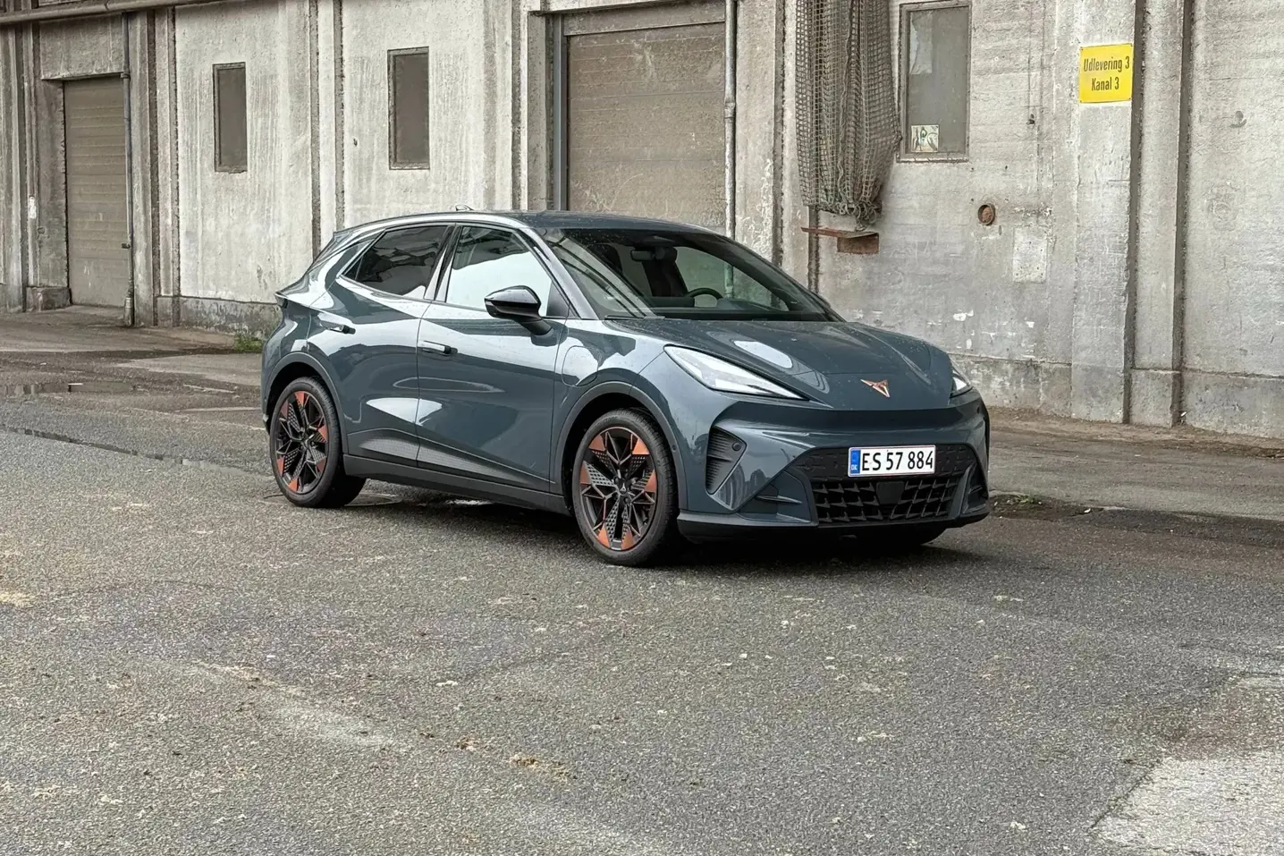 En grå Cupra elbil holder parkeret på en tom, våd industrivej foran en rå betonbygning.