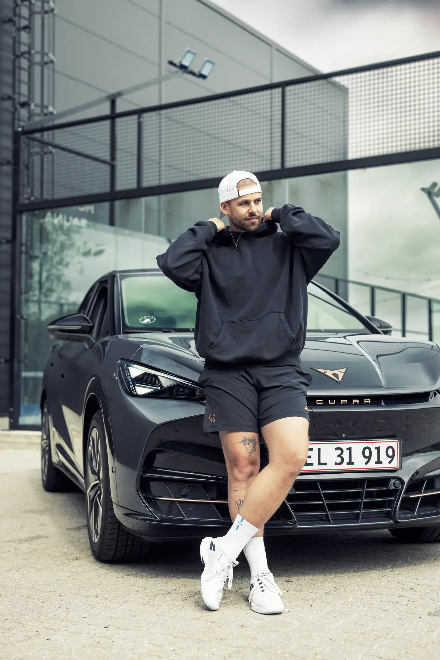Mand i sportstøj læner sig afslappet op ad fronten af en mørkegrå CUPRA-bil foran en moderne bygning.