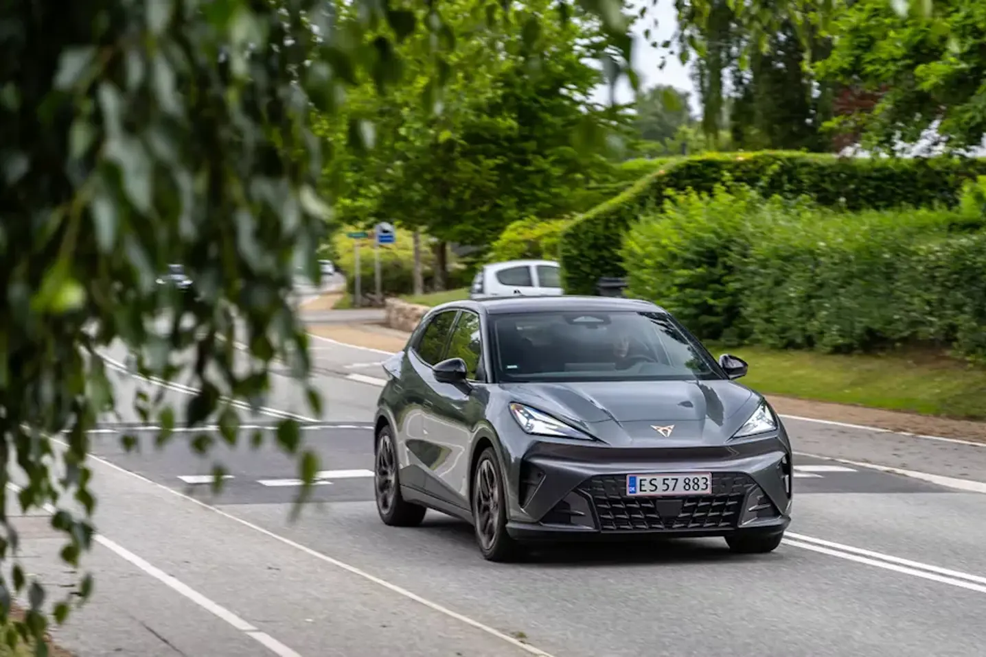 En mørkegrå Cupra elbil kører på en landevej omgivet af grønne træer og hække.