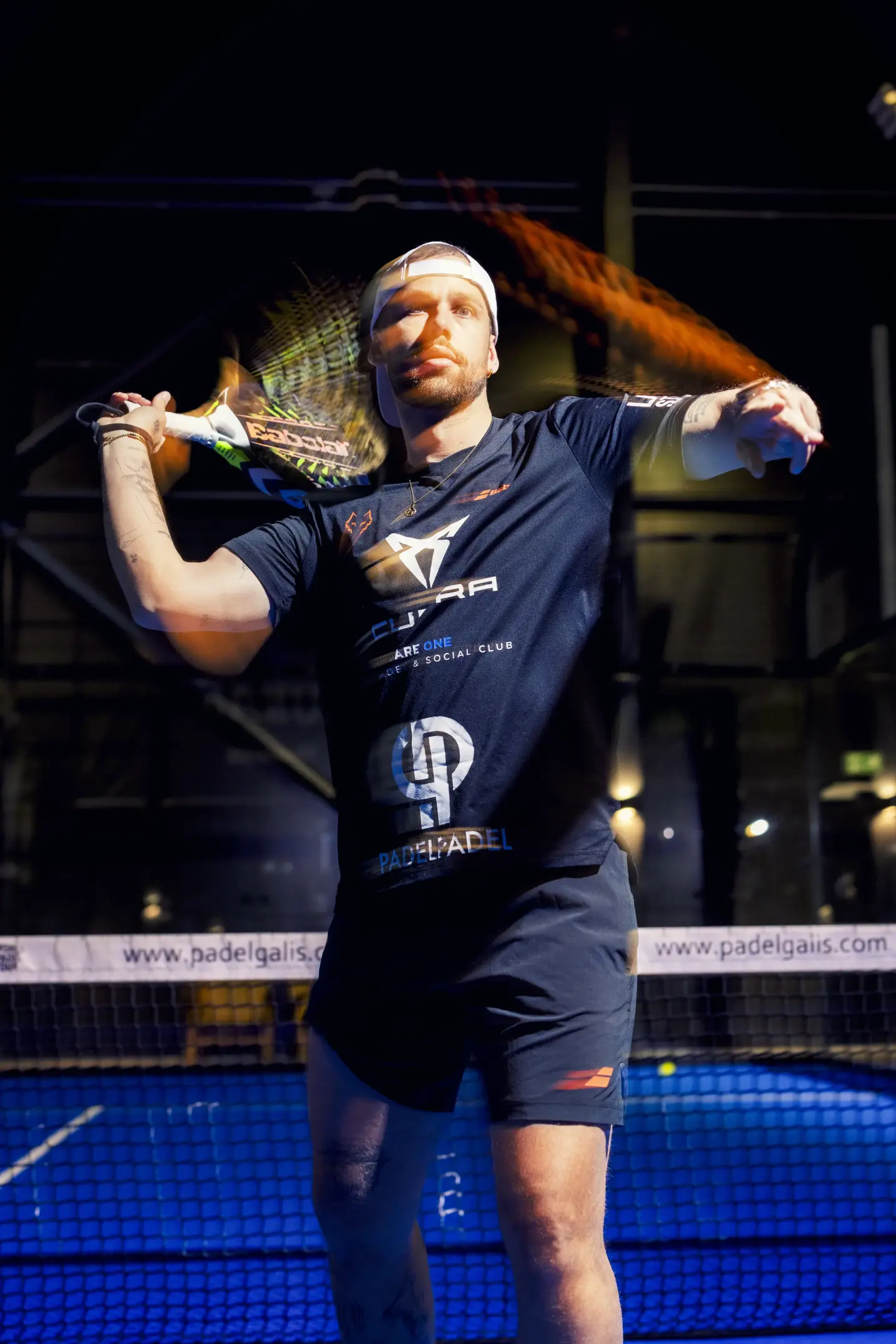 Padelspiller i CUPRA-trøje slår en bold med ketsjeren i et oplyst indendørs padelcenter.