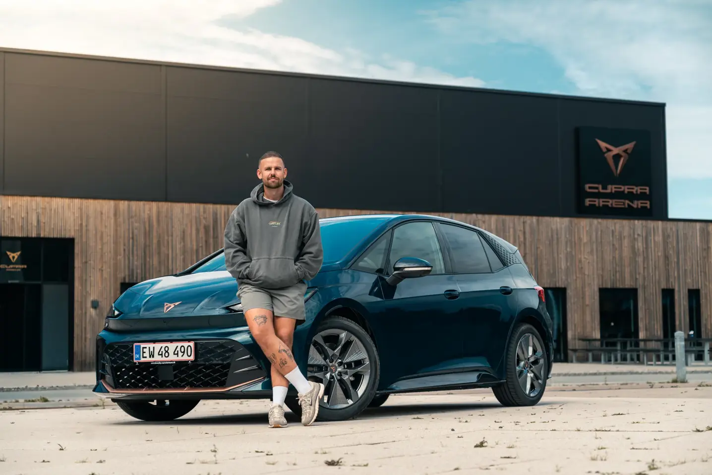 En mand i hoodie læner sig op ad en mørkeblå CUPRA-elbil foran bygningen CUPRA Arena.