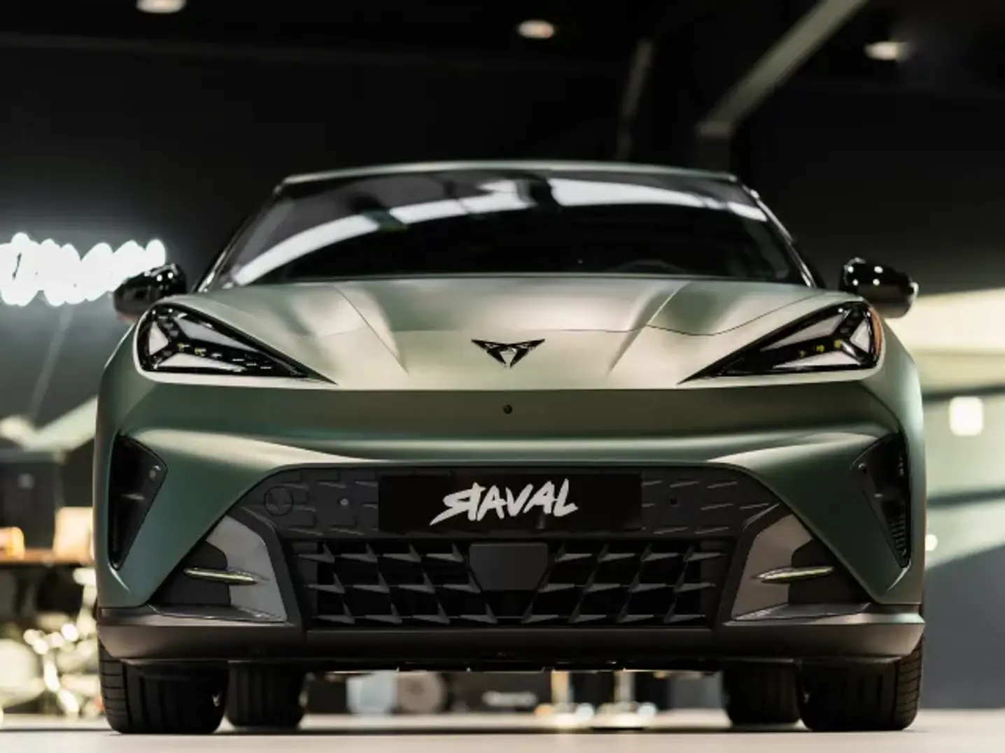 Fronten af en matgrøn, moderne Cupra-bil ses tæt på i et oplyst showroom.