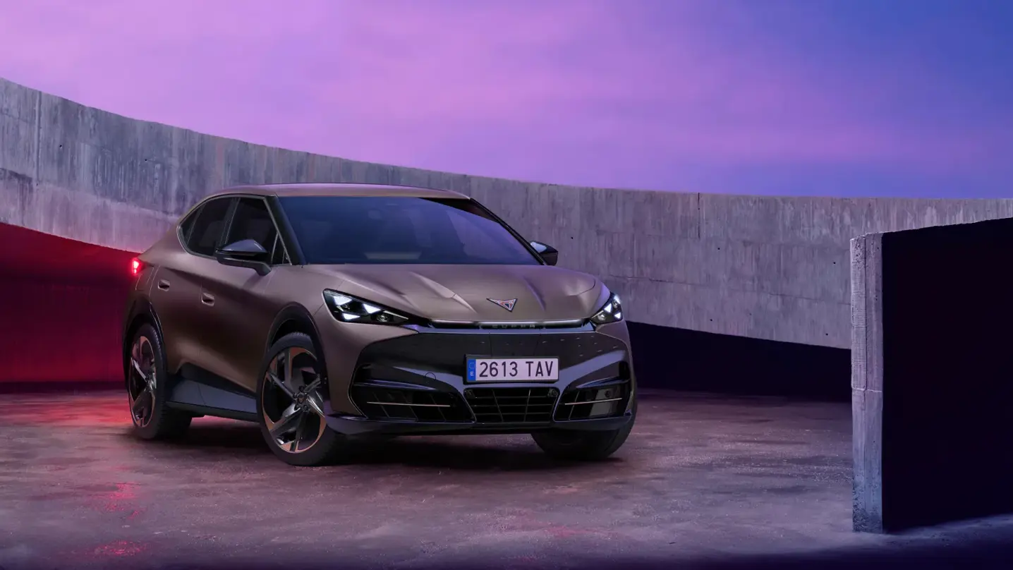 En bronzefarvet Cupra Tavascan elbil holder parkeret i et moderne betonmiljø ved solnedgang med lilla og blå himmel.
