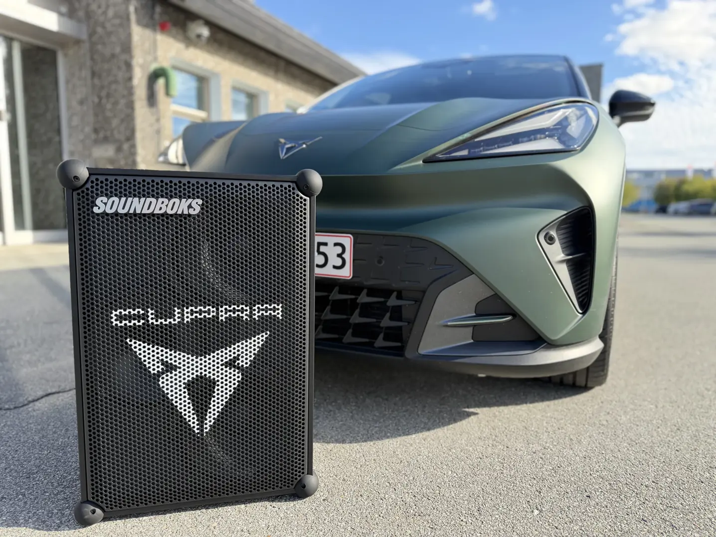 En sort SOUNDOKS-højtaler med CUPRA-logo står på asfalten foran fronten af en grøn CUPRA-bil.